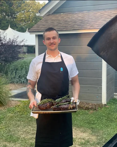 Jake Saari in his apron, holding a tray of roasted meat and herbs outdoors.